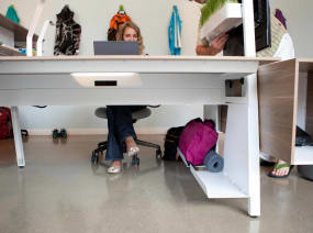 Bivi Bottom Shelf attached to the Bivi Desk frame while a woman works on the desk while a man adjusts a Bivi Planter