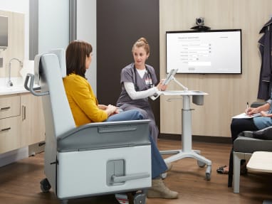 Medical Professional using a tablet with an articulating arm on a Pocket Mobile Workstation while speaking with a patient sitting on an Empath recliner in a examination room