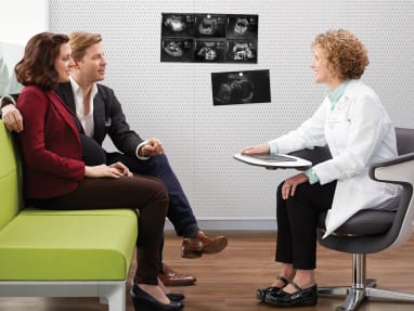 At a hospital room, a profesional talking with a man and woman while seated on a green sofa.