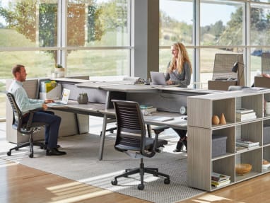 Two people work at a Bivi Dual Height workstation. Steelcase Think desk chairs and Bivi Depot shelving are also pictured.
