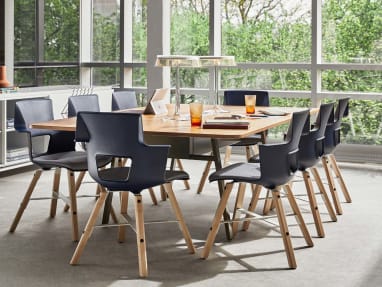 An office meeting area near a window features a collection of Shortcut Wood chairs from Turnstone are arranged around a table. The chairs have a dark blue seat and wooden legs.