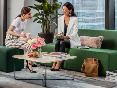 2 business women happily talking, sitting over a green Turnstone Campfire Lounge and a dark-green pouf, with a coffee table in the middle that has books and a flower vase on the top.