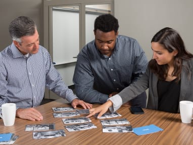 2 men and a woman looking and pointing at photographs laid on top of a wooden table.