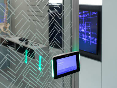 Steelcase Roomwizard scheduling system on a glass wall displays that a conference room is currently available using the screen and LED lights on the side of the tablet