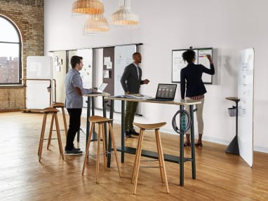 Three people gathered around a Steelcase Roam in a room with Flex standing-height tables and Enea cafe stools