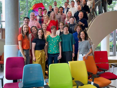 a lot of people posing for the PRIDE month with six office chairs of different colors