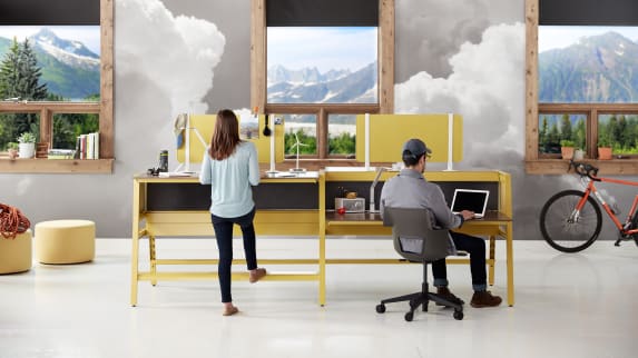 Two people work at a workstation of Bivi desks with yellow legs and privacy screens A woman uses a standing-height worksurface while a man sits in a gray Shortcut 5-Star base chair