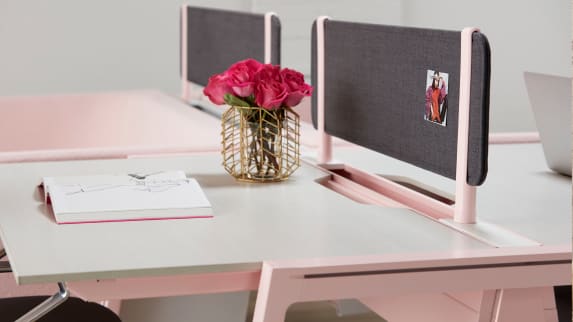 An open book and vase with pink roses is placed on top of a Bivi desk with pink legs and a gray privacy screen
