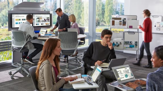 A group of students works together while sitting in Node chairs while another group of people is gathered around a media:scape standing height workstation with cobi chairs next to it