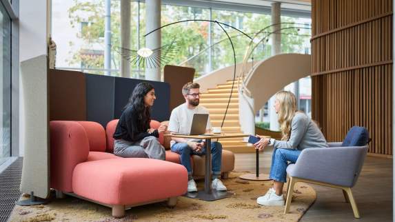 Zona de reuniones informal abierta con una combinación de muebles y colores variados, en un entorno moderno y estéticamente cuidado que fomenta la colaboración.