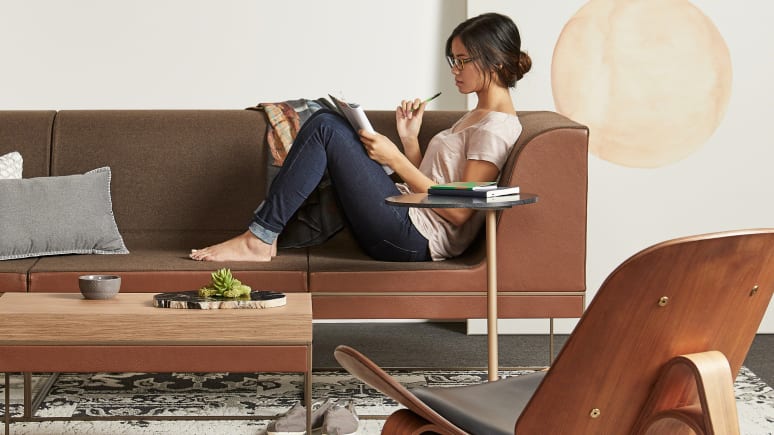 Woman writing on a notebook while seated on a brown Umami sofa with personal table