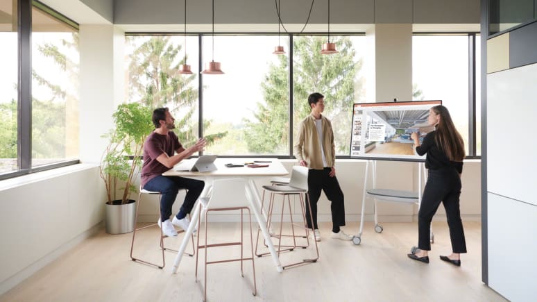 A woman points at a screen displayed on a Steelcase Roam cart close to some Montara stools from and a Potrero415 table from Coalesse