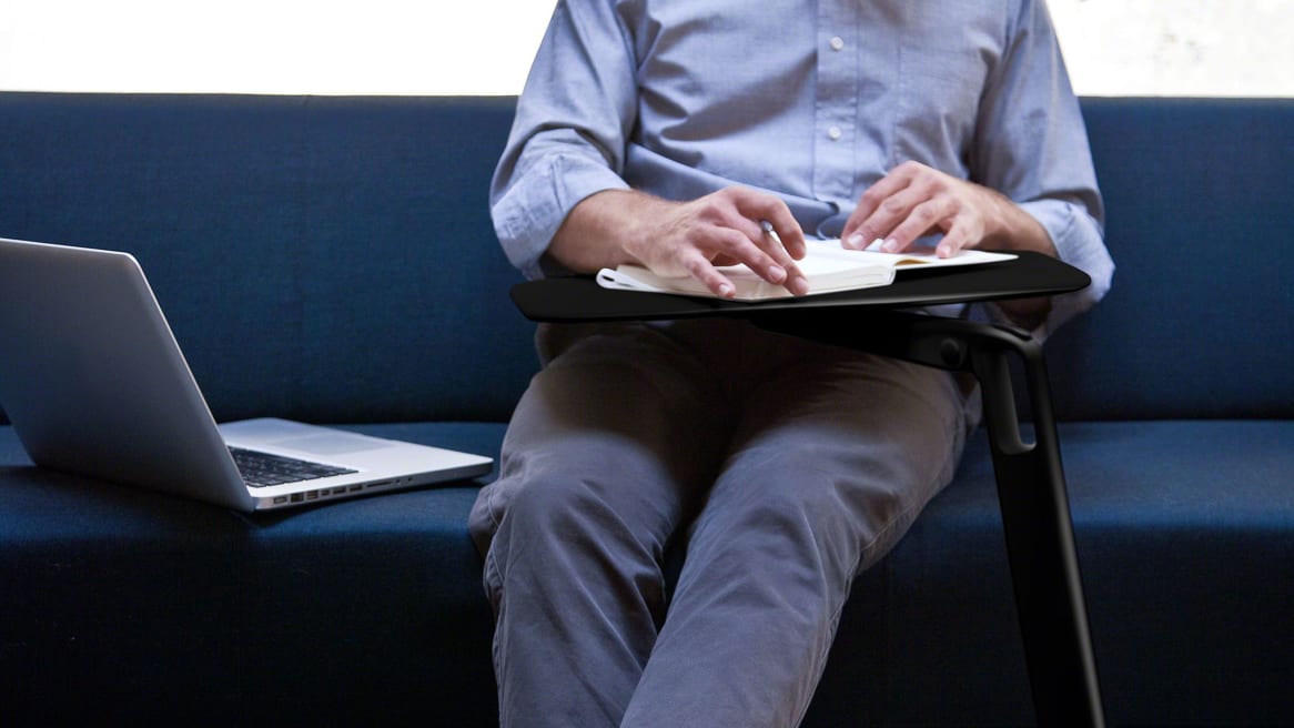 A man holds onto a notebook placed on a Coalesse Free Stand table while a laptop is placed next to him on a sofa