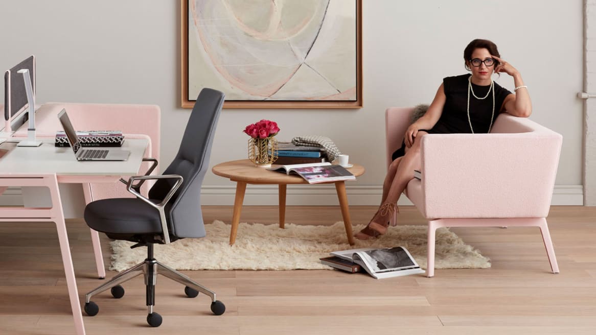 A woman sits on a Bivi Rumble Seat with pink upholstery next to a Bivi Table for Two with pink legs and Coalesse Massaud conference seating with gray upholstery