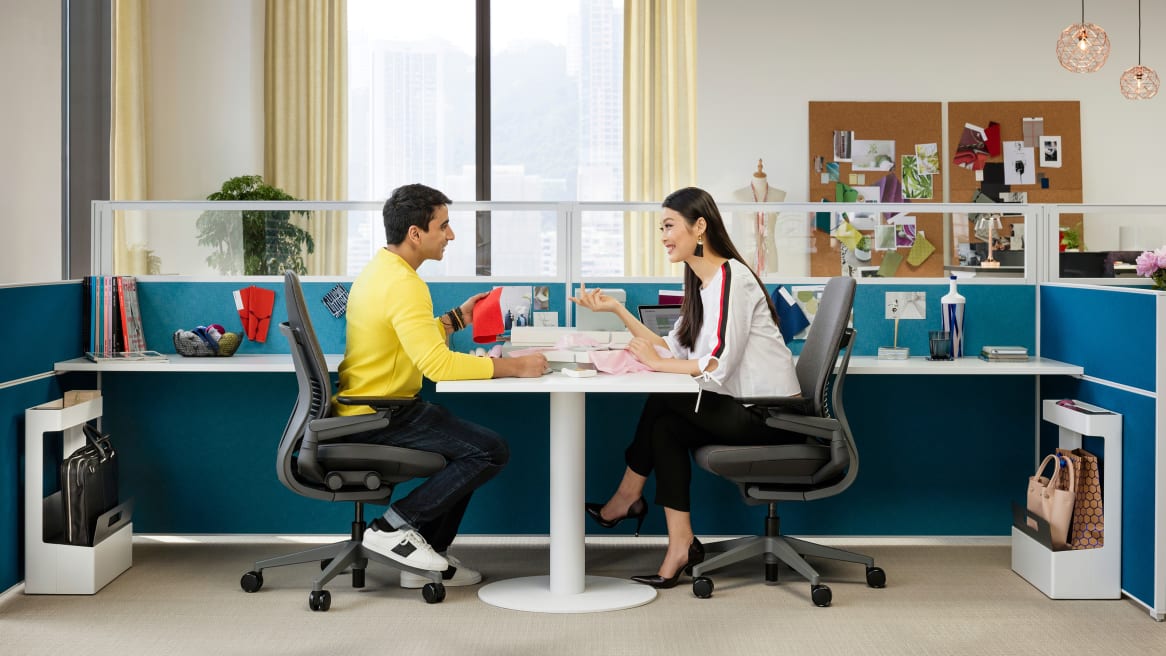 A man and a woman talking over a white desk, while sitting on gray Gesture chairs, surrounded by turquoise wall divisions.