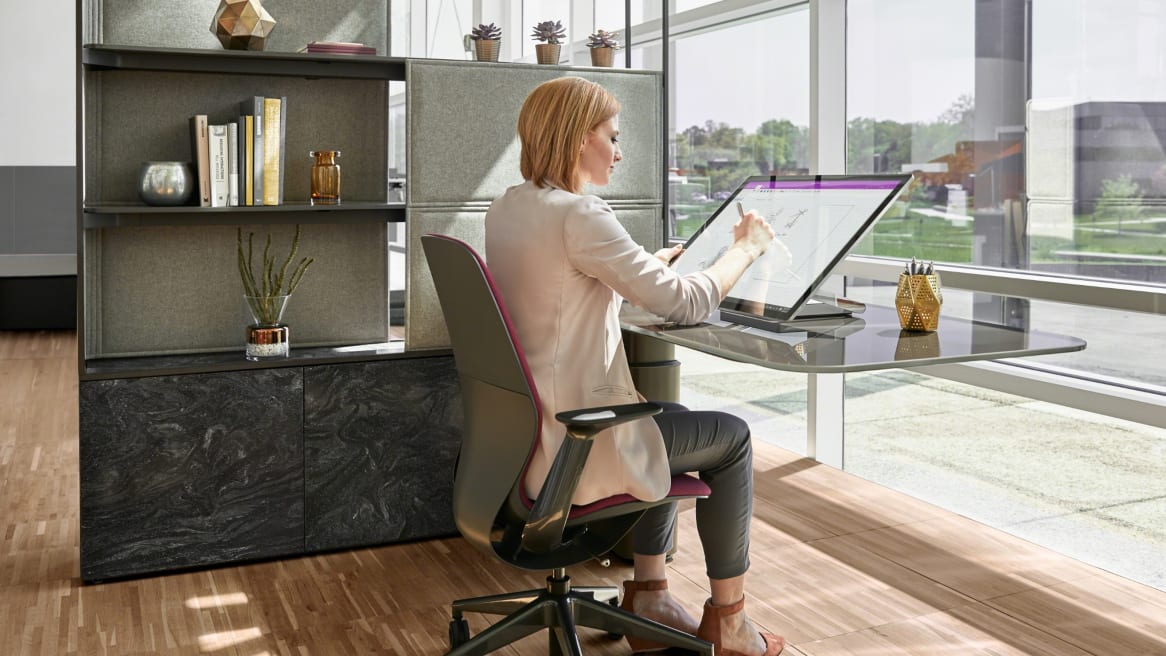 A woman working on Mackinac workstation, sitting on a gray and pink Silq chair while writing on a big tablet.