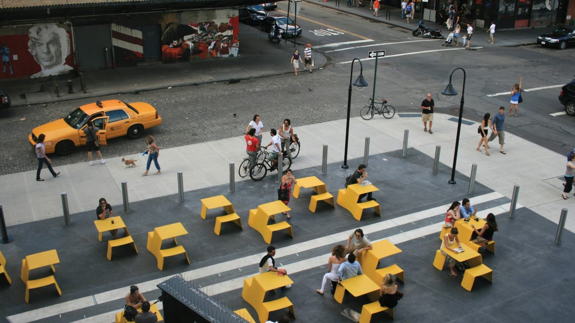 An outdoor space in-between streets with people sitting and talking on yellow Extremis Picnik Table.