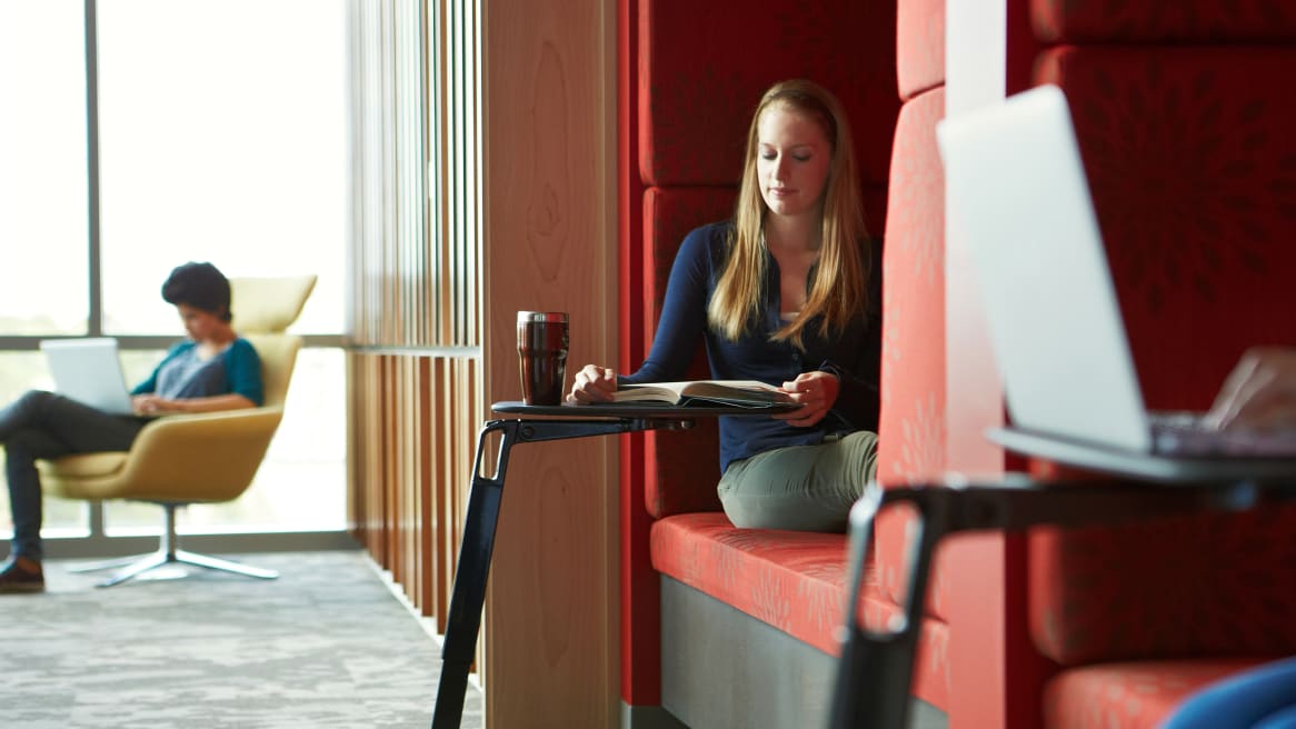 A woman reading a book while she sits on a red lounge booth with a personal table.