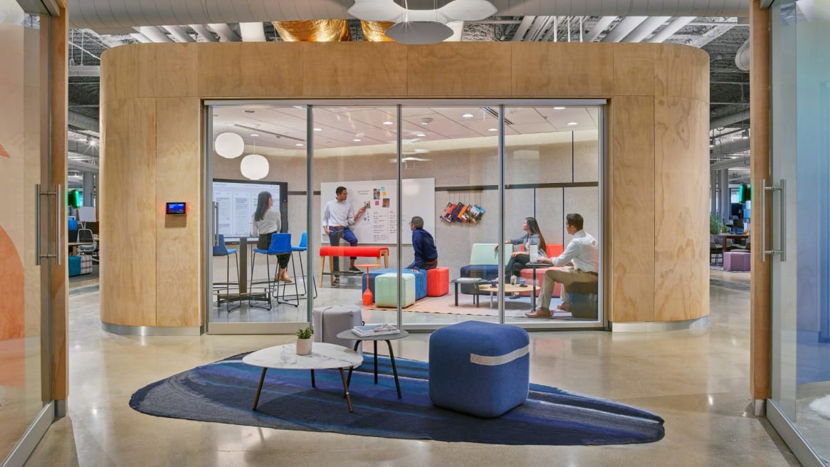in between space with two ottomans and two tables in front of a large project space with glass and wood walls where a team is looking at a whiteboard