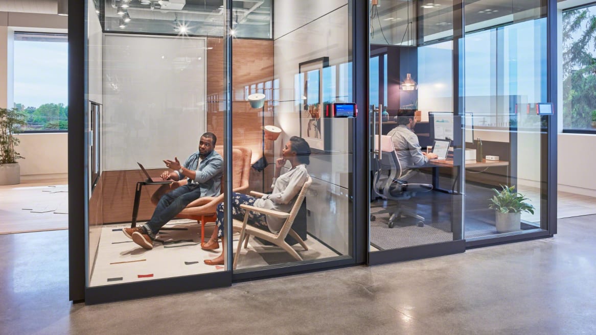 In one meeting room, a woman sits in a CH25 Paddle Chair with another person while a man works while sitting in a Steelcase Gesture chair in a neighboring enclave