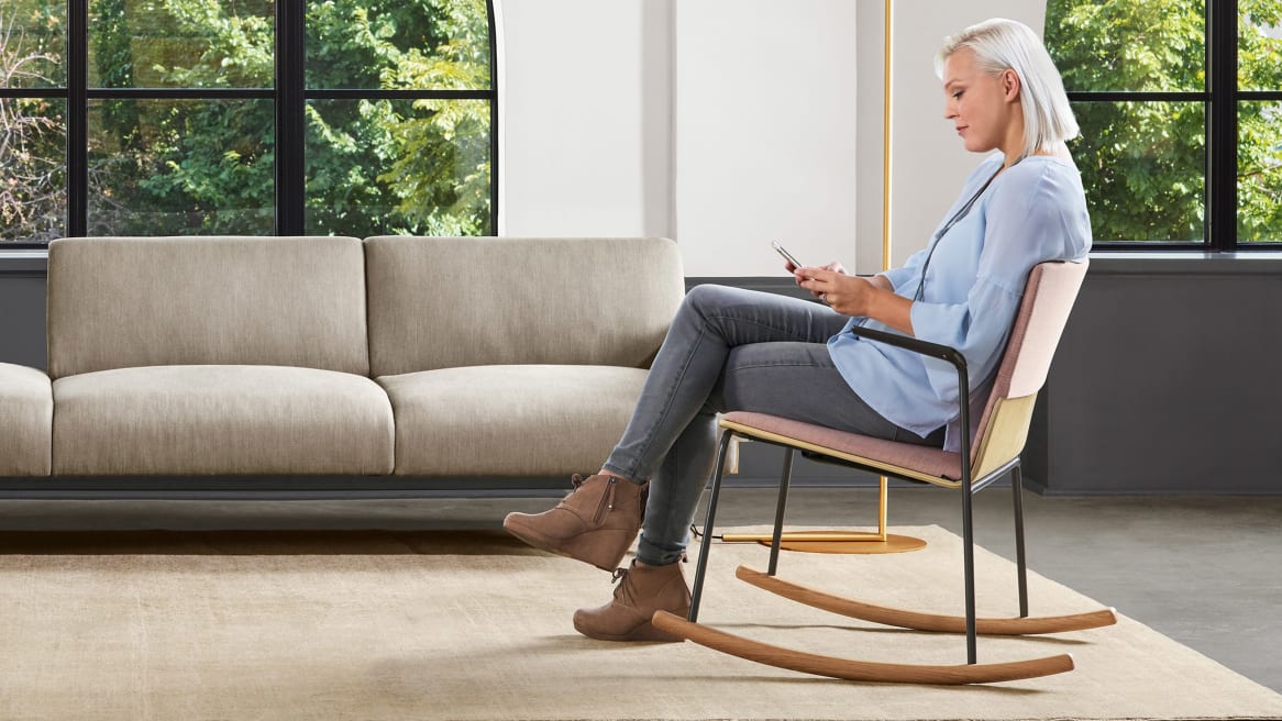 A woman sits in a Montara650 rocker chair while looking at her phone in an office lounge. A Sistema lounge system is also pictured.