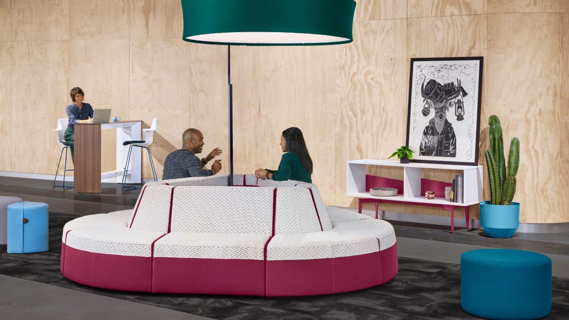 Two people have a conversation while sitting on a Campfire Lounge System in an island configuration Bassline Credenza, Campfire Ottoman, Campfire Pouf, Campfire Slim standing table, Campfire Big Lamp, and Shortcut X-Base stools are also pictured