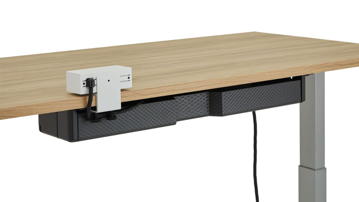 The back of a black Steelcase Universal Cable Management Kit attached under a light wood height-adjustable desk.