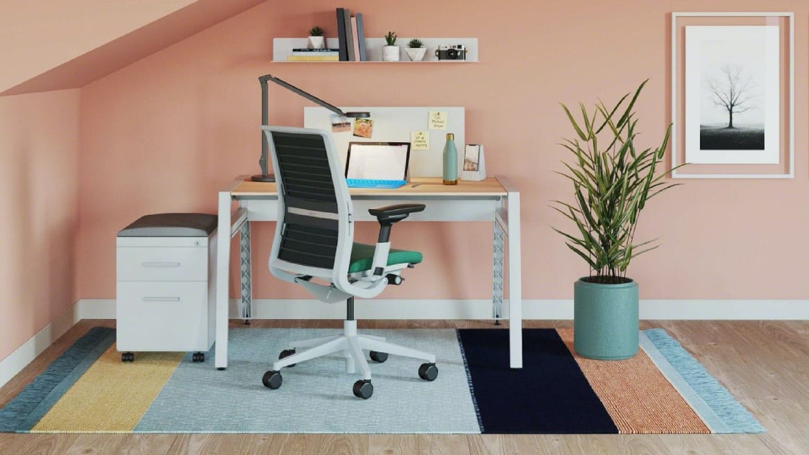 Private home office with a pink wall, wooden desk with a laptop on it, a plant and a blue Think chair
