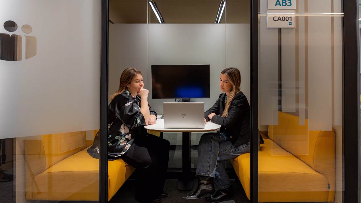Two people seated at a small table with a laptop inside a glass-enclosed meeting room