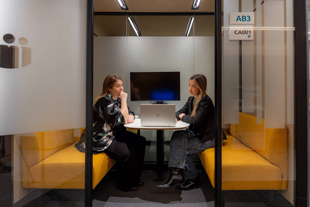 Two people seated at a small table with a laptop inside a glass-enclosed meeting room
