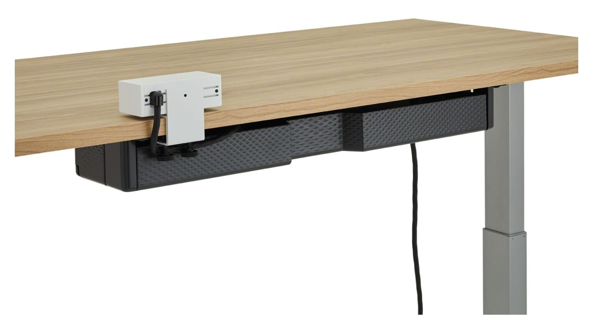 The back of a black Steelcase Universal Cable Management Kit attached under a light wood height-adjustable desk.